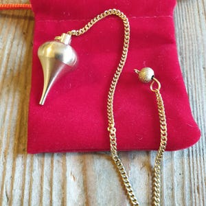 Könnte beinhalten: Ein goldfarbenes Metallpendel mit einem glatten, tropfenförmigen Gewicht, das an einer feinen Gliederkette befestigt ist. Die Kette hat am Ende eine kleine, texturierte, kugelförmige Perle. Das Pendel liegt auf einem roten Samtbeutel.