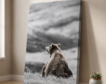Könnte beinhalten: Ein rechteckiger Leinwanddruck mit einer Schwarz-Weiß-Fotografie eines Bären, der von hinten gesehen in einem grasbewachsenen Feld sitzt. Der Bär befindet sich unten in der Mitte, blickt nach links und ist vor einer unscharfen Hügellandschaft.