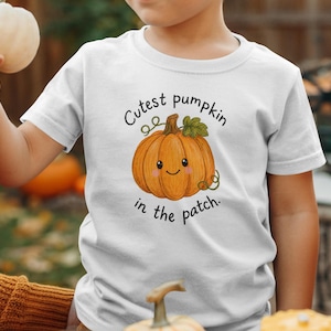 Puede incluir: Una camiseta blanca de manga corta con un gráfico de una calabaza naranja sonriente con hojas y enredaderas verdes. El texto "Cutest pumpkin in the patch." está impreso en letras negras sobre y debajo de la ilustración.