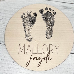 May include: A round wooden plaque with two black baby footprints and the names "Mallory Jayde" written in black and brown lettering.