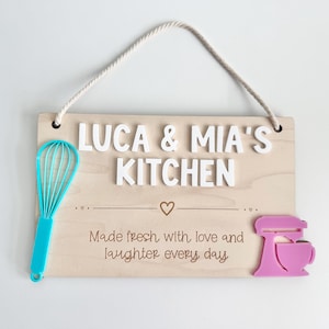 The image shows a wooden sign with a rope for hanging. It reads "LUCA & MIA'S KITCHEN" with "Made fresh with love and laughter every day" There's a turquoise whisk on the left and a pink mixer on the right. It’s intended use is play kitchen decor