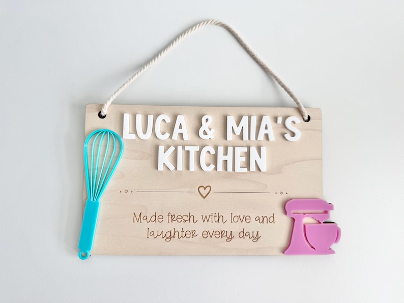 The image shows a wooden sign with a rope for hanging. It reads "LUCA & MIA'S KITCHEN" with "Made fresh with love and laughter every day" There's a turquoise whisk on the left and a pink mixer on the right. It’s intended use is play kitchen decor