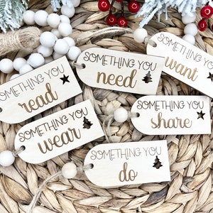 Könnte beinhalten: Geschenkanhänger aus Holz mit den Aufschriften „Something You Read“, „Something You Wear“, „Something You Need“, „Something You Want“, „Something You Share“ und „Something You Do“. Jedes Etikett hat ein Stern- oder Baumdesign. Die Etiketten sind mit Schnur und Holzperlen aufgereiht.