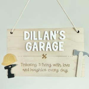 May include: Wooden sign with the words "DILLAN'S GARAGE" in white, 3D letters. The sign includes a yellow hard hat, a black drill, a gray hammer, and the text "Tinkering & fixing with love and laughter every day."