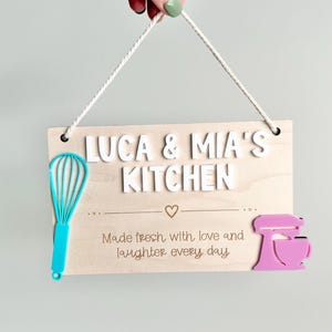 The image shows a wooden sign with a rope for hanging. It reads "LUCA & MIA'S KITCHEN" with "Made fresh with love and laughter every day" There's a turquoise whisk on the left and a pink mixer on the right. Playroom Decor
