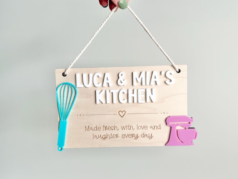 The image shows a wooden sign with a rope for hanging. It reads "LUCA & MIA'S KITCHEN" with "Made fresh with love and laughter every day" There's a turquoise whisk on the left and a pink mixer on the right. Playroom Decor