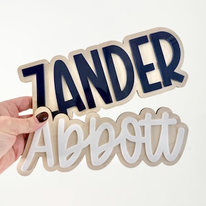 Könnte beinhalten: Zwei Namensschilder aus Holz. Eines zeigt den Namen "ZANDER" in Marineblau, das andere "Abbott" in Weiß. Beide Namen sind auf einem hellen Holzgrund übereinander angeordnet.