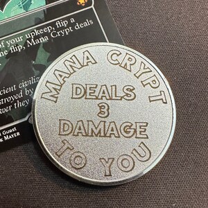 May include: A silver-colored metal token with the words "Mana Crypt Deals 3 Damage To You" printed in brown lettering.