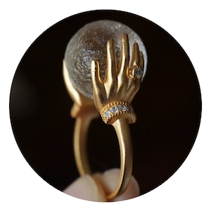 May include: Gold ring featuring two hands holding a clear glass orb. The hands have a diamond ring on one finger and a band of small diamonds around the wrist.