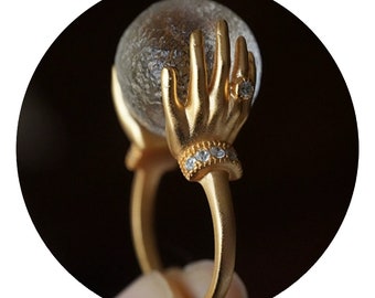 May include: Gold ring featuring two hands holding a clear glass orb. The hands have a diamond ring on one finger and a band of small diamonds around the wrist.