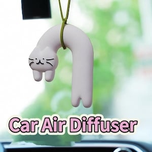 Könnte beinhalten: Ein weißer Keramik-Lufterfrischer für das Auto in Form einer Katze mit eingerolltem Schwanz. Die Katze hat schwarze Augen und eine schwarze Nase. Der Lufterfrischer hängt an einer grünen Schnur.