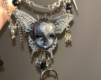 May include: Silver-toned keychain featuring a butterfly-faced pendant with intricate details. The pendant has a face with a butterfly wing design, accented with black beads and a small bat charm. The keychain includes a chain and a key ring.