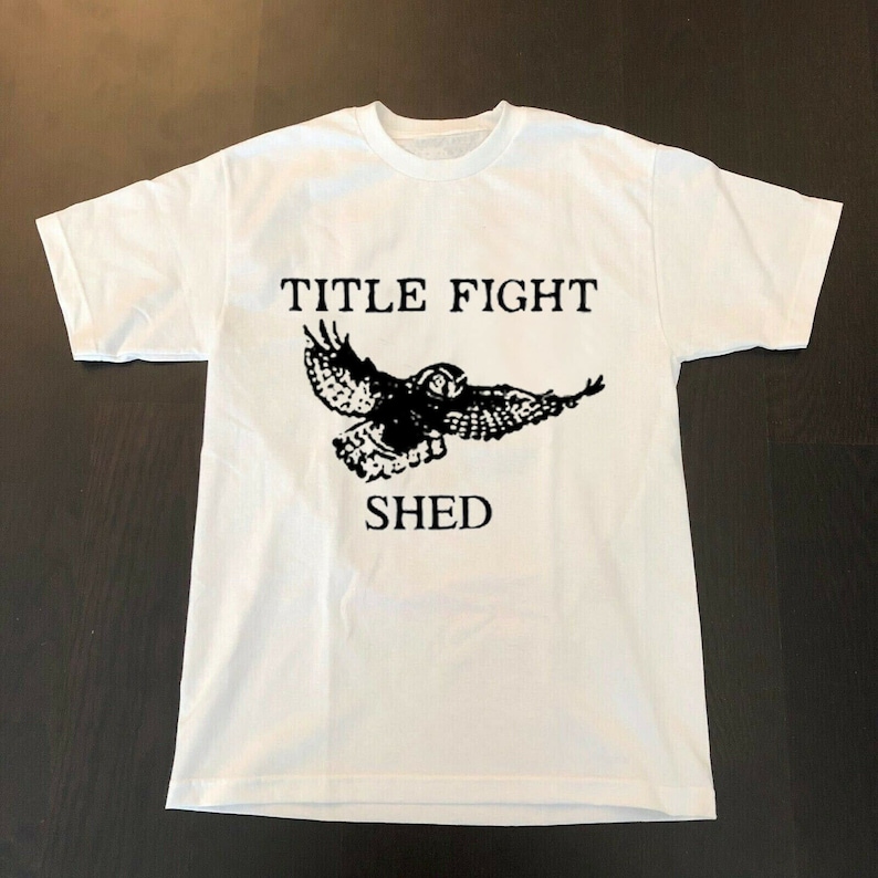 以下が含まれることがあります： 白いTシャツに、飛んでいるフクロウの黒いグラフィックと「TITLE FIGHT SHED」の文字がプリントされています。