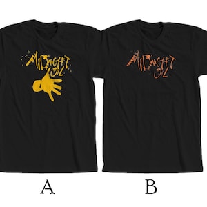以下が含まれることがあります： 前面にバンド名「Midnight Oil」がプリントされた黒いTシャツ2枚。左側のTシャツは黄色の手形、右側のTシャツはオレンジ色の手形がプリントされています。