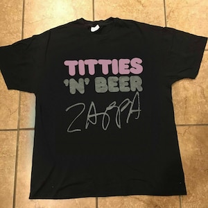 Puede incluir: Camiseta negra con un gráfico rosa y gris que dice "TITTIES 'N' BEER ZAPPA".