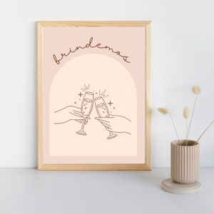 May include: A printable art print with a peach background and a brown outline of two champagne flutes clinking together. The text "brindemos" is written in a brown cursive font above the flutes.