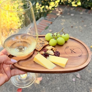 May include: A wooden serving board with a cutout for a wine glass stem, featuring a laser-engraved letter K inside a circular wreath design. The board holds two slices of yellow cheese, a small pile of mixed nuts, and a cluster of green grapes.