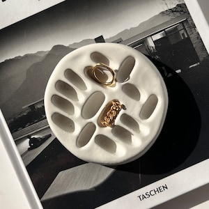 Könnte beinhalten: Eine weiße Keramik-Ring-Schale mit mehreren Vertiefungen zum Aufbewahren von Schmuck. Die Schale liegt auf einem schwarz-weißen Magazin mit dem Titel "NEUTRA BARBARA LAMPRECHT TASCHEN".