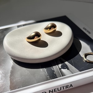 Puede incluir: Un par de pendientes de forma de lágrima de color dorado sobre una superficie de piedra blanca. Los pendientes son pequeños y delicados.