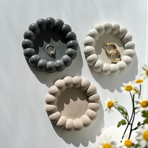 May include: Three round concrete trinket dishes with a raised, textured edge. The dishes are in shades of grey, white, and beige. The grey dish has a gold ring inside. The white dish has a pair of gold hoop earrings inside.