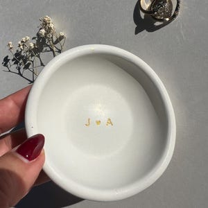 May include: A white ceramic ring dish with gold lettering of the initials "J & A" and a heart symbol. Two gold rings with a diamond are on the upper right. A small sprig of dried flowers is on the left.