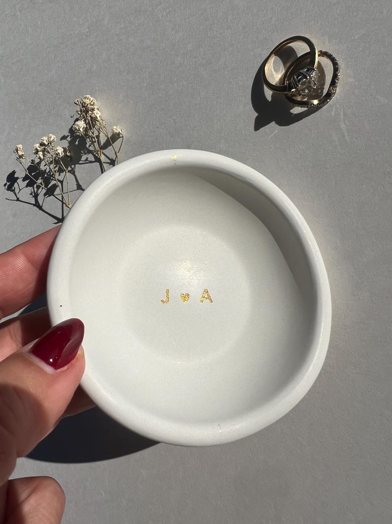 May include: A white ceramic ring dish with gold lettering of the initials "J & A" and a heart symbol. Two gold rings with a diamond are on the upper right. A small sprig of dried flowers is on the left.