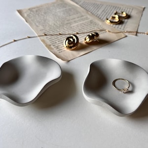 Peut inclure: Deux plats en c&eacute;ramique blanche aux bords ondul&eacute;s. Un plat contient une bague en or avec une petite pierre. L'autre plat est vide.