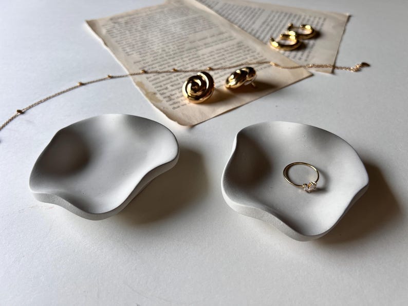 Peut inclure: Deux plats en c&eacute;ramique blanche aux bords ondul&eacute;s. Un plat contient une bague en or avec une petite pierre. L'autre plat est vide.