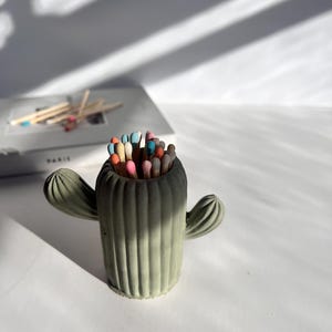 May include: A green concrete cactus shaped matchstick holder with colourful matchsticks inside.