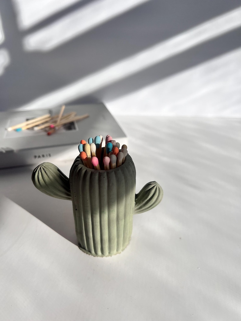 May include: A green concrete cactus shaped matchstick holder with colourful matchsticks inside.