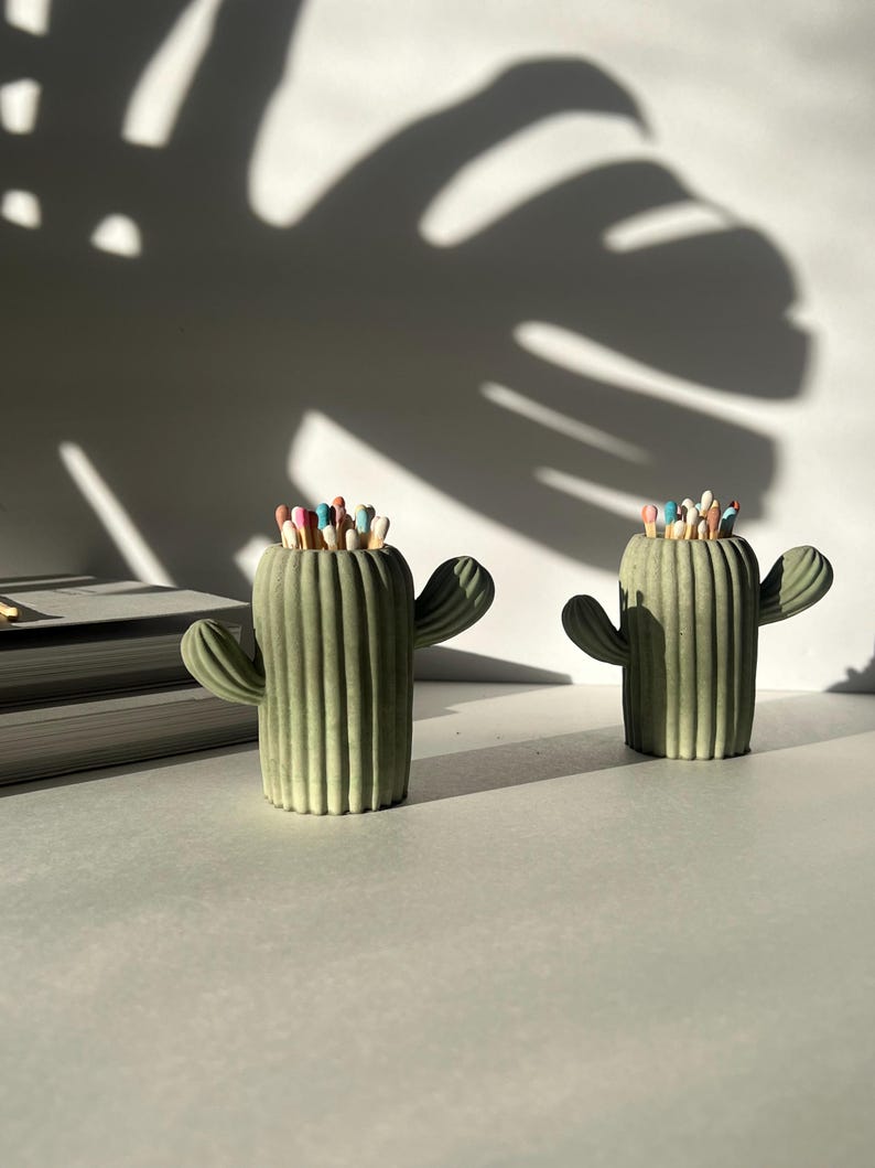 May include: Two green cactus shaped matchstick holders with colourful matchsticks inside. The holders are on a white surface.