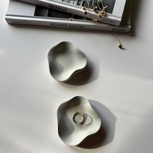 Peut inclure: Deux coupelles &agrave; anneaux en c&eacute;ramique blanche avec une forme ondul&eacute;e et organique. Les coupelles sont sur une surface blanche. Une coupelle contient une bague en or.