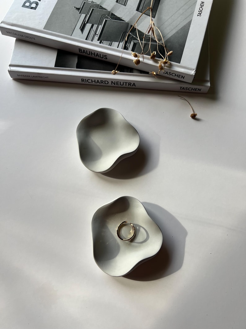 Peut inclure: Deux coupelles &agrave; anneaux en c&eacute;ramique blanche avec une forme ondul&eacute;e et organique. Les coupelles sont sur une surface blanche. Une coupelle contient une bague en or.