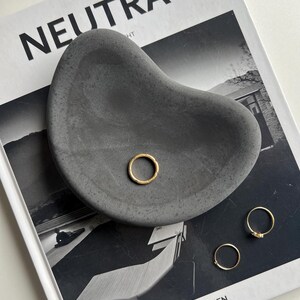 K&ouml;nnte beinhalten: Eine dunkelgraue, organisch geformte Betonschale mit drei goldenen Ringen. Die Schale liegt auf einem Schwarz-Wei&szlig;-Magazin mit dem Wort "NEUTRA" auf dem Cover.