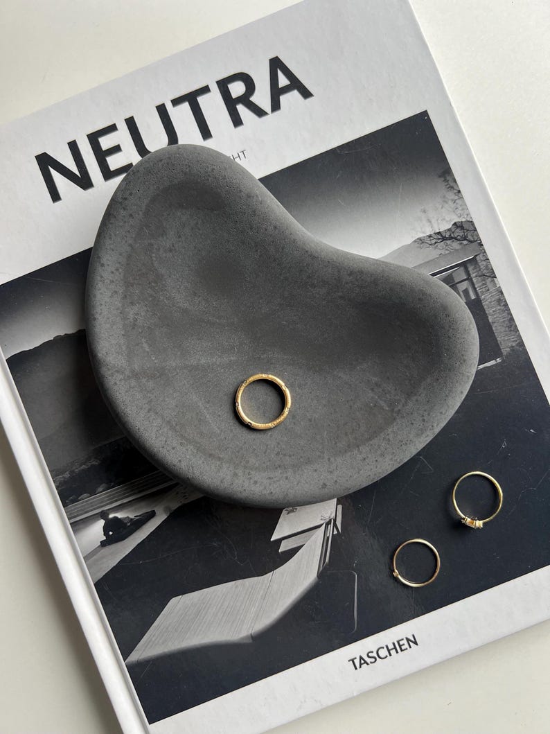 K&ouml;nnte beinhalten: Eine dunkelgraue, organisch geformte Betonschale mit drei goldenen Ringen. Die Schale liegt auf einem Schwarz-Wei&szlig;-Magazin mit dem Wort "NEUTRA" auf dem Cover.