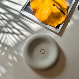 May include: A light gray ceramic dish with a ring resting in the center. The dish is on a white surface with a shadow of a plant leaf.