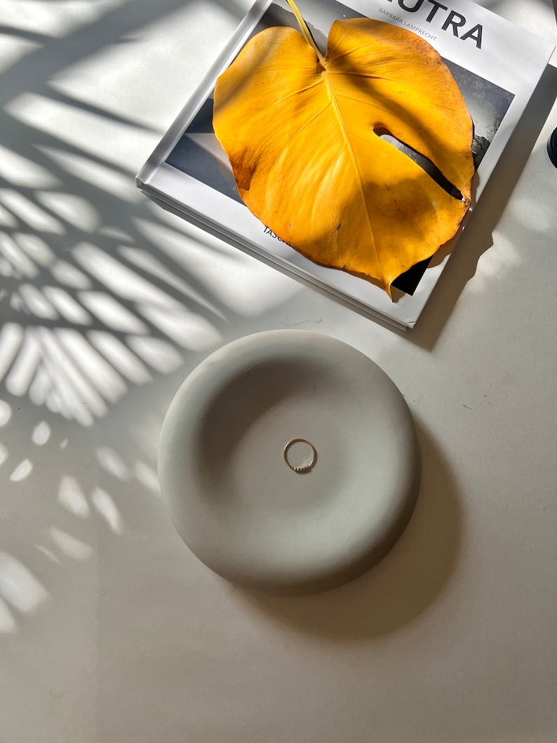May include: A light gray ceramic dish with a ring resting in the center. The dish is on a white surface with a shadow of a plant leaf.
