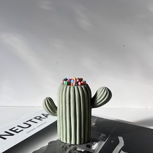 May include: A green concrete cactus shaped pencil holder with colourful pencils inside. The cactus has a ribbed texture and two arms.