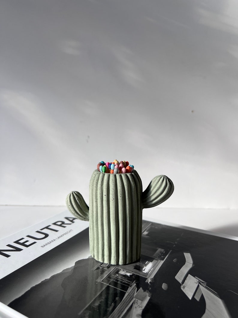 May include: A green concrete cactus shaped pencil holder with colourful pencils inside. The cactus has a ribbed texture and two arms.