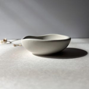 May include: A small, off-white ceramic bowl with a gently curved rim. The bowl has a smooth, matte finish and casts a soft shadow. The design is simple and elegant, suitable for holding small items like jewelry or trinkets.
