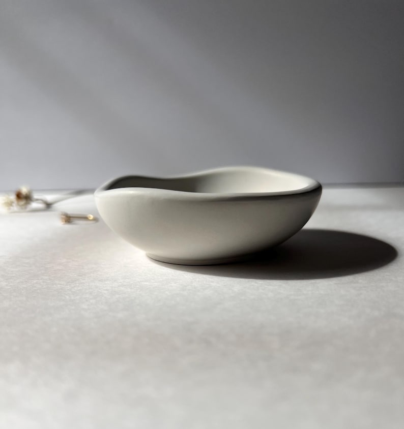 May include: A small, off-white ceramic bowl with a gently curved rim. The bowl has a smooth, matte finish and casts a soft shadow. The design is simple and elegant, suitable for holding small items like jewelry or trinkets.