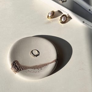 Peut inclure: Un présentoir à bagues rond en céramique blanc cassé avec un accent marron texturé. Une bague en or avec un petit diamant est posée sur le présentoir. Boucles d'oreilles dorées et autres bagues sur un livre avec "Paris" et "London".