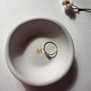 May include: A small, round, white ceramic dish with the gold letter "M" in the center. A delicate gold ring with small stones rests inside the dish. A sprig of dried white flowers is in the background.