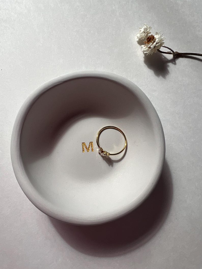 May include: A small, round, white ceramic dish with the gold letter "M" in the center. A delicate gold ring with small stones rests inside the dish. A sprig of dried white flowers is in the background.