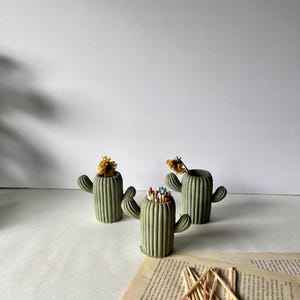 May include: Three green concrete cactus shaped match holders. One holder is filled with colourful matches. The holders are sitting on a white surface with a book underneath.