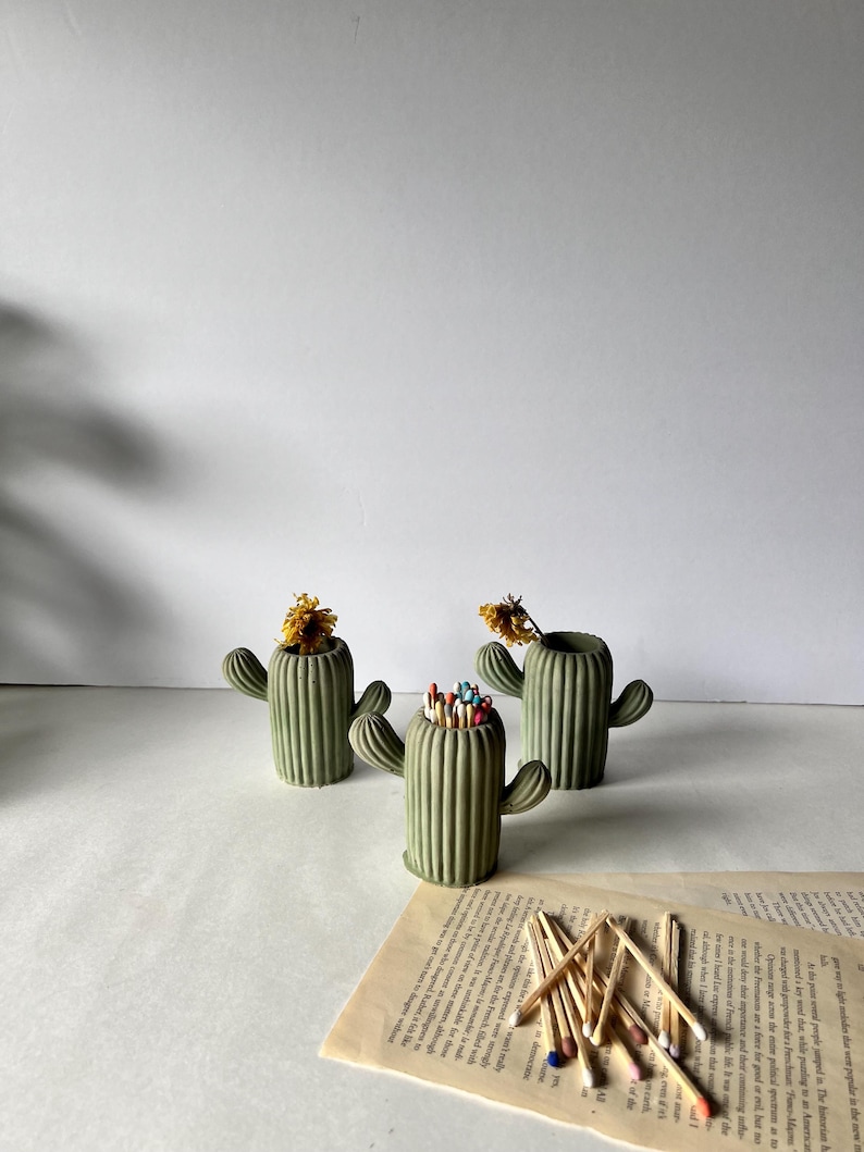 May include: Three green concrete cactus shaped match holders. One holder is filled with colourful matches. The holders are sitting on a white surface with a book underneath.
