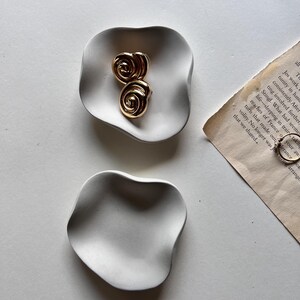 Peut inclure: Deux plats en c&eacute;ramique blanche avec une forme ondul&eacute;e et organique. Le plus grand plat a deux boucles d'oreilles dor&eacute;es en forme de spirale sur le dessus.