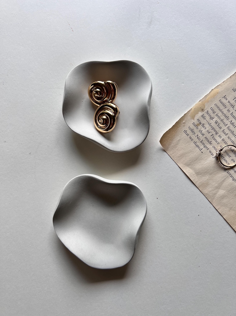 Peut inclure: Deux plats en c&eacute;ramique blanche avec une forme ondul&eacute;e et organique. Le plus grand plat a deux boucles d'oreilles dor&eacute;es en forme de spirale sur le dessus.