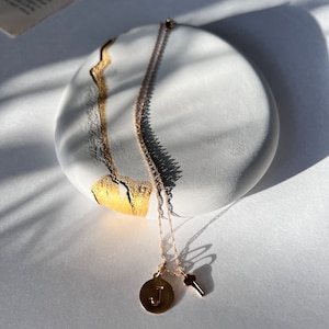 May include: A gold necklace with a key charm and a circular pendant with the letter "J" on a white and gold speckled oval-shaped dish.