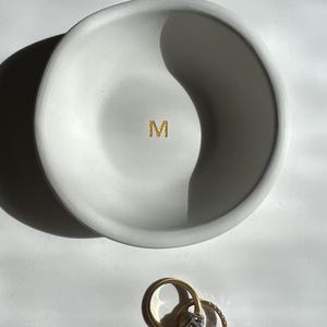 May include: A white, round, shallow dish with a gold glitter "M" in the center. Two gold rings with gemstones are next to the dish. The dish is likely a jewelry holder.